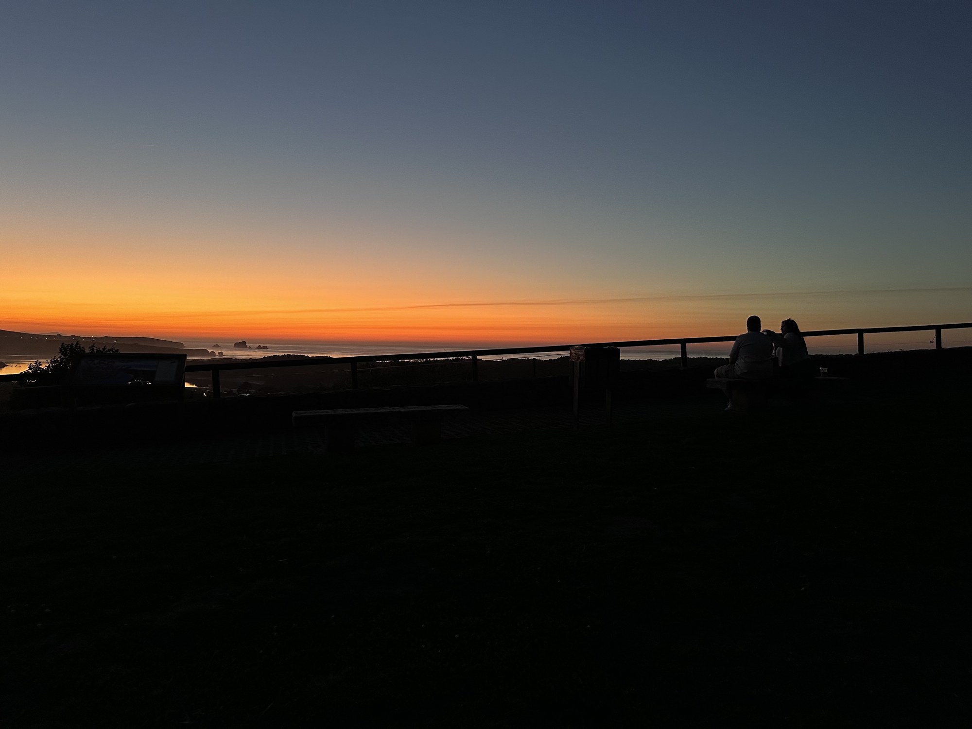 Two people sitting on a bench at a coastal lookout, watching the sunset over the sea.